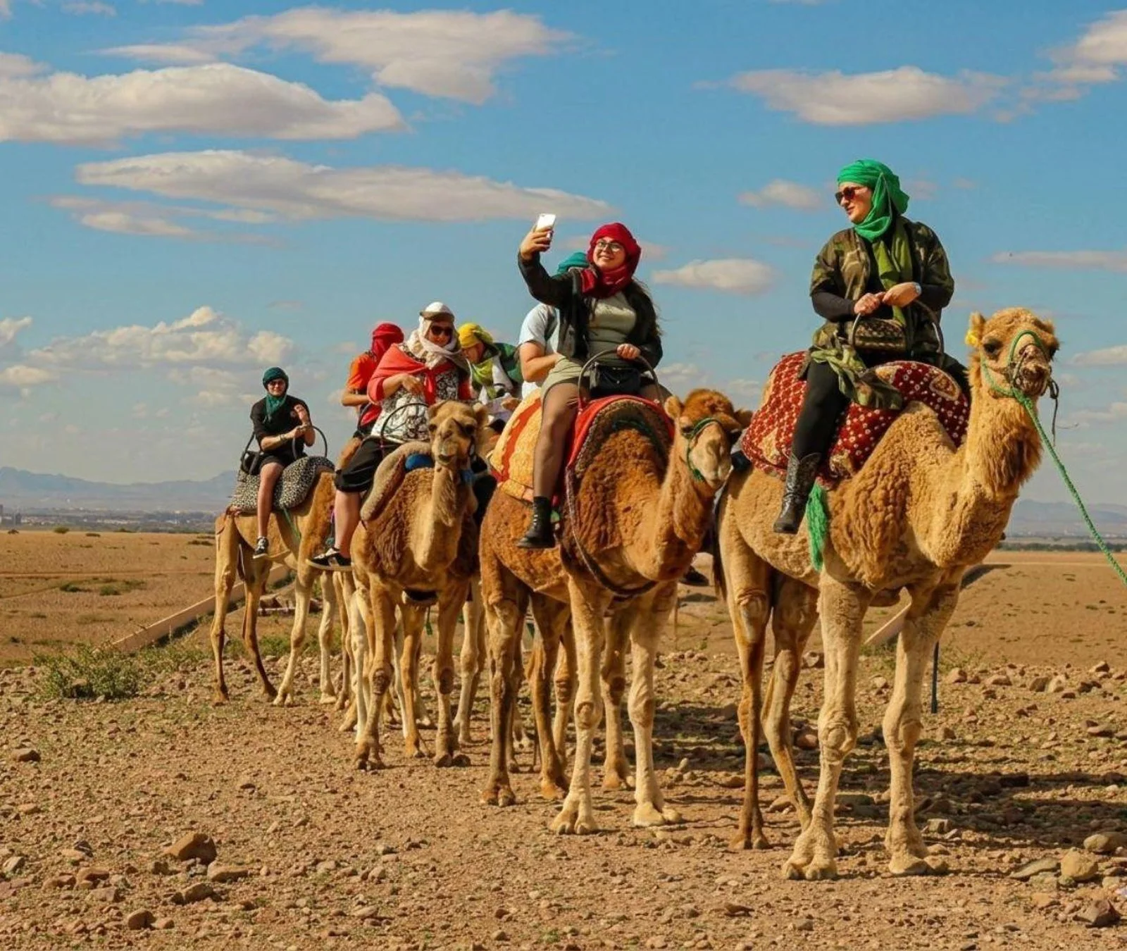 Tourists riding a line of camels across the rocky Agafay Desert, one taking a selfie